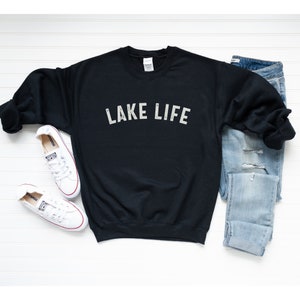 May include: Black sweatshirt with the text "LAKE LIFE" in white on the front. The sweatshirt is laid flat on a white surface with a pair of white sneakers and blue jeans.