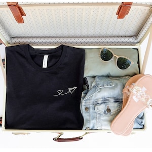 以下が含まれることがあります： 旅行に必要な物が詰まった開いたスーツケース。黒いTシャツには紙飛行機とハートのグラフィックがプリントされています。サングラス、デニムジャケット、ピンクのサンダルも入っています。