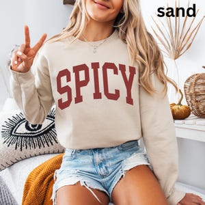 Może przedstawiać: Kobieta w jasnobeżowej bluzie z nadrukiem "SPICY" w kolorze bordowym z przodu. Ma na sobie dżinsowe spodenki i rozpuszczone włosy.