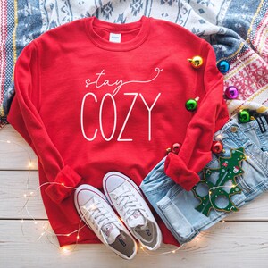 May include: Red sweatshirt with the text "stay cozy" in white lettering. The sweatshirt is lying on a white wooden surface with a pair of white trainers, blue jeans, and a green Christmas tree bauble.