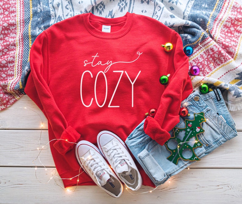 May include: Red sweatshirt with the text "stay cozy" in white lettering. The sweatshirt is lying on a white wooden surface with a pair of white trainers, blue jeans, and a green Christmas tree bauble.