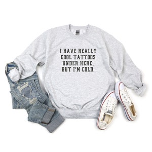 May include: A gray sweatshirt with the text "I have really cool tattoos under here, but I'm cold." printed on the front. The sweatshirt is paired with blue jeans and white sneakers.