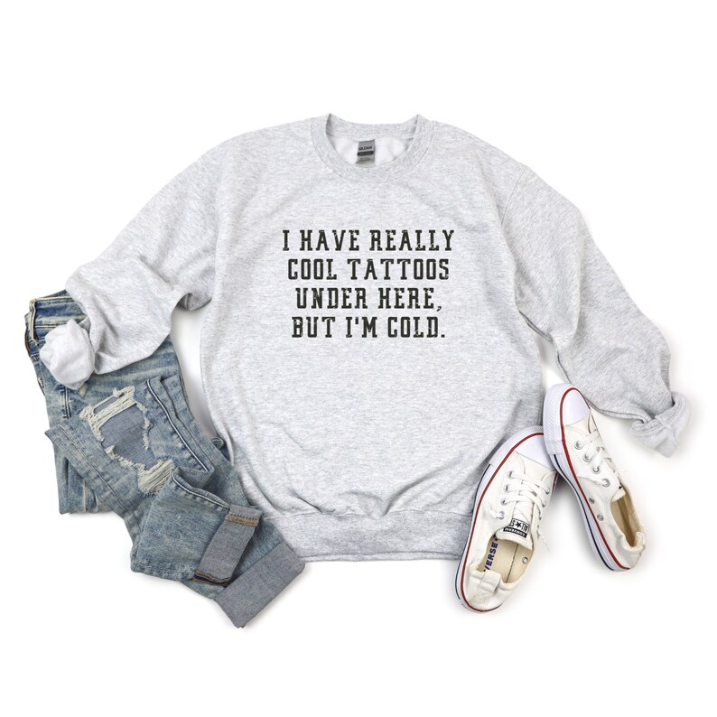 May include: A gray sweatshirt with the text "I have really cool tattoos under here, but I'm cold." printed on the front. The sweatshirt is paired with blue jeans and white sneakers.