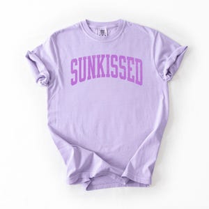 May include: A light purple t-shirt with the word "SUNKISSED" printed in a darker purple, arched font across the chest. The shirt has short sleeves and a crew neck.