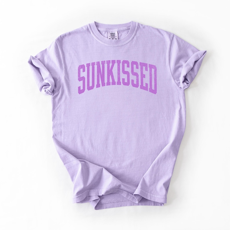 May include: A light purple t-shirt with the word "SUNKISSED" printed in a darker purple, arched font across the chest. The shirt has short sleeves and a crew neck.