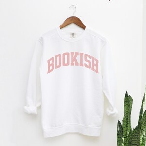 Puede incluir: Sudadera blanca con un gr&aacute;fico rosa 'BOOKISH' en una fuente universitaria.