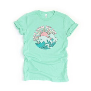 Op de afbeelding: Een lichtgroen T-shirt met een retro-grafische afbeelding van een zon die opkomt boven een golf. De afbeelding zegt "Here Comes The Sun".