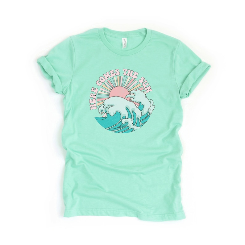 Op de afbeelding: Een lichtgroen T-shirt met een retro-grafische afbeelding van een zon die opkomt boven een golf. De afbeelding zegt "Here Comes The Sun".