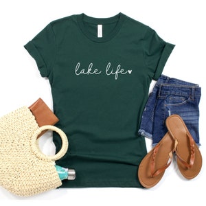 K&ouml;nnte beinhalten: Ein dunkelgr&uuml;nes T-Shirt mit dem Text "lake life" in wei&szlig;er Schrift mit einem kleinen Herzsymbol. Das Shirt liegt flach auf einer wei&szlig;en Oberfl&auml;che mit einer Strohtasche, einer braunen Brieftasche, einer Wasserflasche und einem Paar braunen Sandalen.