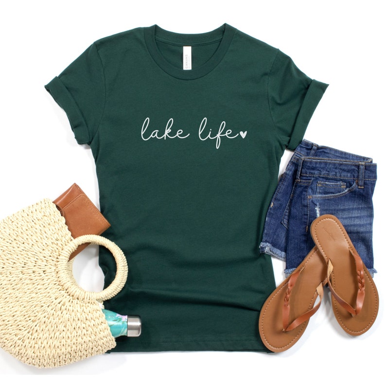K&ouml;nnte beinhalten: Ein dunkelgr&uuml;nes T-Shirt mit dem Text "lake life" in wei&szlig;er Schrift mit einem kleinen Herzsymbol. Das Shirt liegt flach auf einer wei&szlig;en Oberfl&auml;che mit einer Strohtasche, einer braunen Brieftasche, einer Wasserflasche und einem Paar braunen Sandalen.