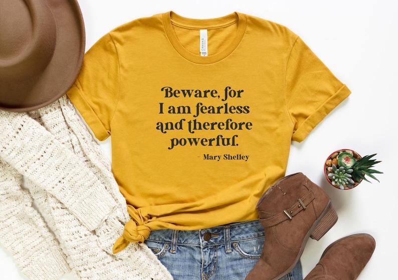 Puede incluir: Una camiseta amarilla mostaza con una cita negra que dice "Beware, for I am fearless and therefore powerful. - Mary Shelley".