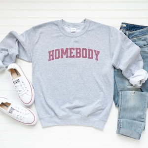 Puede incluir: Una sudadera gris con la palabra "HOMEBODY" impresa en burdeos en la parte delantera. La sudadera se combina con unos vaqueros azules y zapatillas blancas.