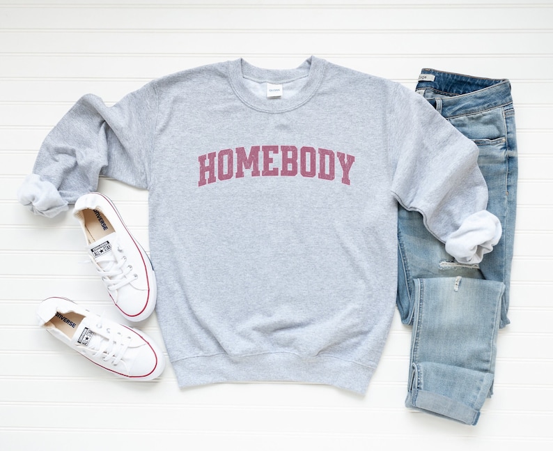 Puede incluir: Una sudadera gris con la palabra "HOMEBODY" impresa en burdeos en la parte delantera. La sudadera se combina con unos vaqueros azules y zapatillas blancas.
