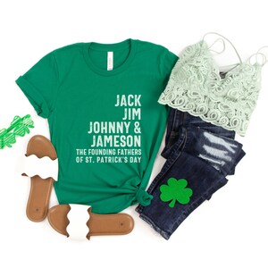 May include: Green t-shirt with white text that reads "Jack Jim Johnny & Jameson The Founding Fathers of St. Patrick's Day". The shirt is paired with a green lace top, blue jeans with a green shamrock patch, and white sandals.
