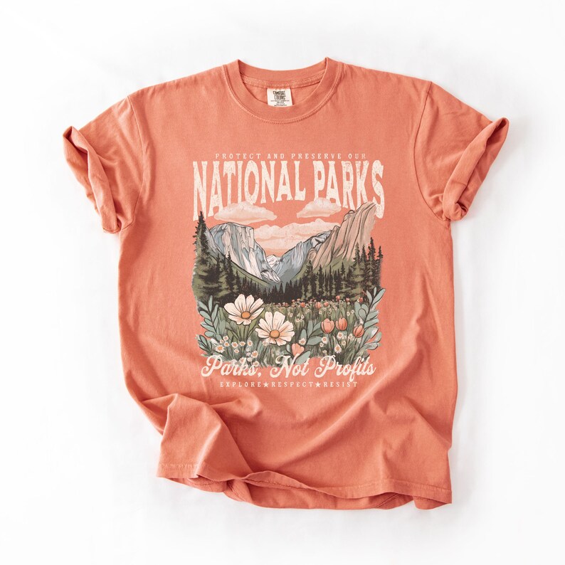 K&ouml;nnte beinhalten: Ein orangefarbenes T-Shirt mit einem grafischen Aufdruck einer Berglandschaft mit Wald und Blumen. Der Text "Protect and Preserve Our National Parks" befindet sich &uuml;ber dem Bild und der Text "Parks, Not Profits" unter dem Bild. Der Text "Explore, Respect, Resist" befindet sich unter dem Text "Parks, Not Profits".