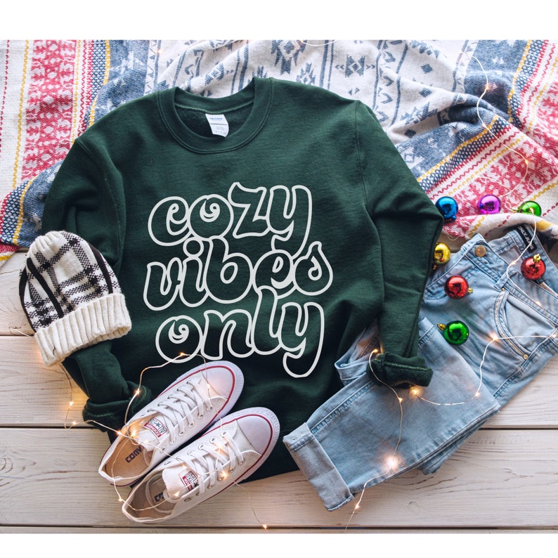 May include: A green sweatshirt with white text that reads "cozy vibes only". The sweatshirt is laid out on a white wooden surface with a pair of white sneakers, blue jeans, and a plaid beanie.