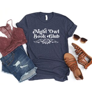 May include: A navy blue t-shirt with the text "Night Owl Book Club" in white, featuring a stylized owl graphic. The shirt is displayed on a white background with a pair of brown sandals, a denim skirt, and a lace bra.