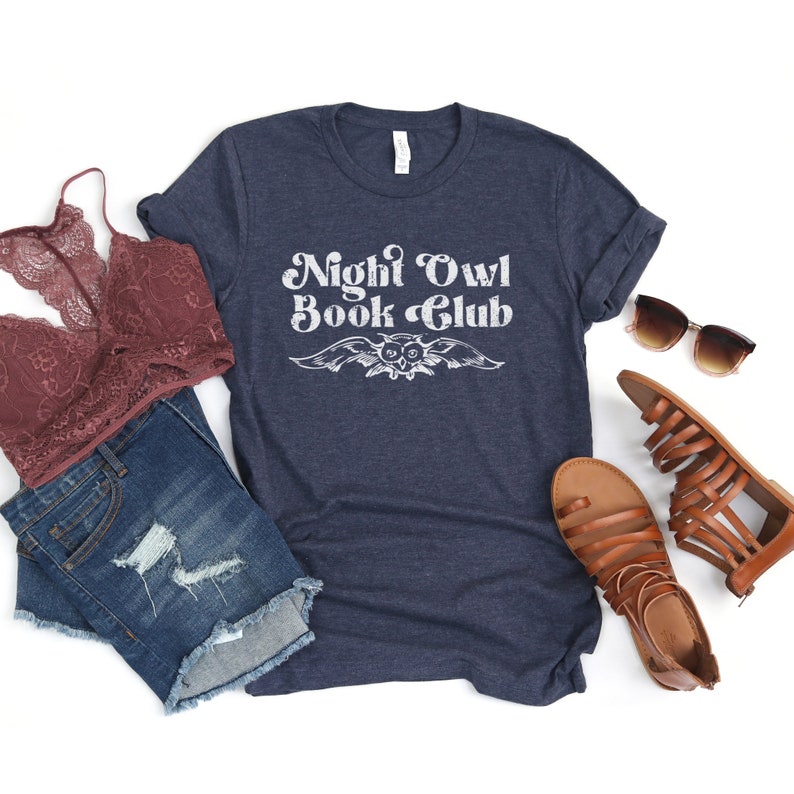 May include: A navy blue t-shirt with the text "Night Owl Book Club" in white, featuring a stylized owl graphic. The shirt is displayed on a white background with a pair of brown sandals, a denim skirt, and a lace bra.