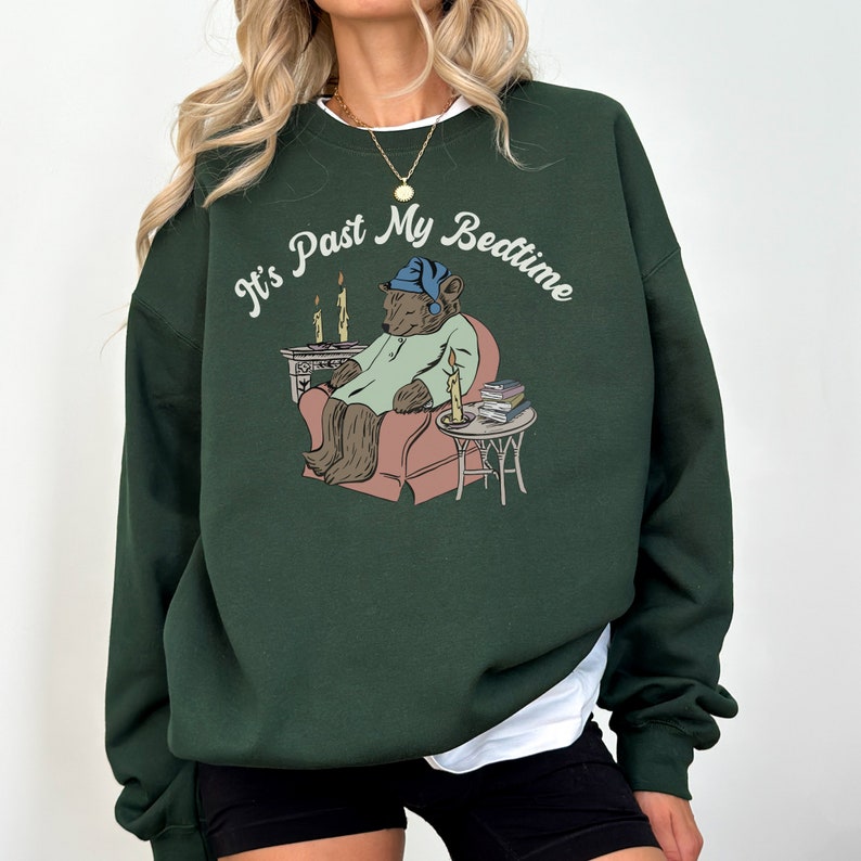 Pu&ograve; includere: Una felpa verde scuro con una grafica di un orso che indossa un berretto da notte e un pigiama, seduto su una sedia con un libro e delle candele. Il testo "It's Past My Bedtime" &egrave; stampato in un font corsivo bianco.