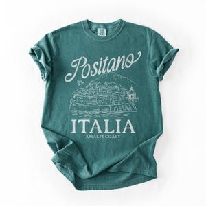 May include: Teal-colored t-shirt with the word "Positano" in a script font above a white line drawing of a coastal town. The word "ITALIA" is printed below the illustration, with "AMALFI COAST" underneath.