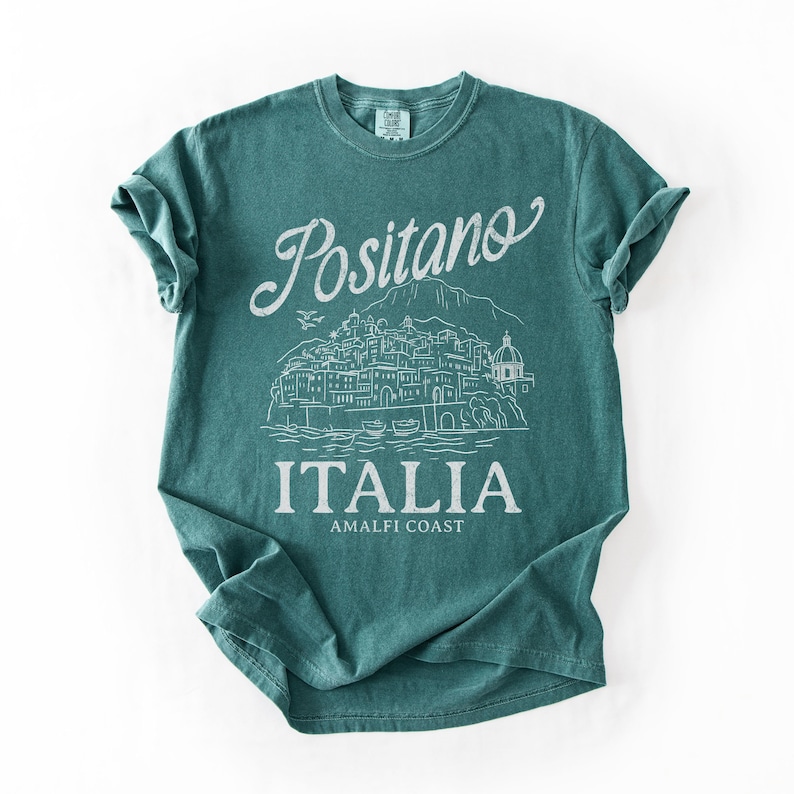 May include: Teal-colored t-shirt with the word "Positano" in a script font above a white line drawing of a coastal town. The word "ITALIA" is printed below the illustration, with "AMALFI COAST" underneath.