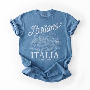 May include: A blue t-shirt with a white graphic of the town of Positano, Italy. The text "Positano" is arched above the illustration, with "ITALIA" and "AMALFI COAST" below. The shirt is made of soft material.