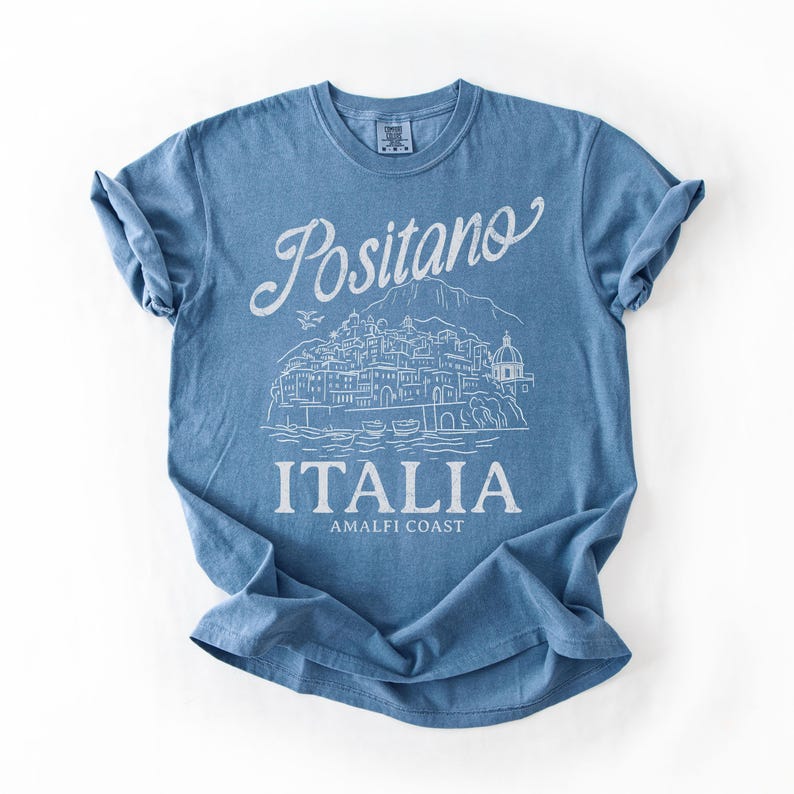 May include: A blue t-shirt with a white graphic of the town of Positano, Italy. The text "Positano" is arched above the illustration, with "ITALIA" and "AMALFI COAST" below. The shirt is made of soft material.
