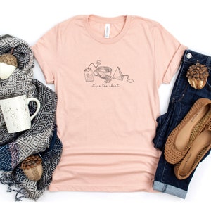 May include: A peach-colored t-shirt with a graphic of a teacup, tea bag, and tea infuser. The text "it's a tea shirt" is printed below the graphic.