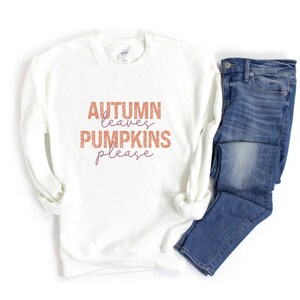 以下が含まれることがあります： オレンジと紫の文字で「AUTUMN leaves PUMPKINS please」と書かれた白いスウェットシャツ。カジュアルな秋の装いとして、青いデニムジーンズと組み合わせています。
