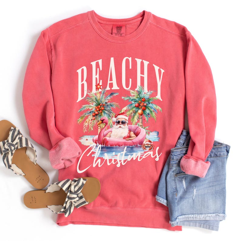 May include: Coral-colored sweatshirt with the words "BEACHY Christmas" and a graphic of Santa Claus in sunglasses floating on a flamingo floatie. The image includes denim shorts and striped sandals.