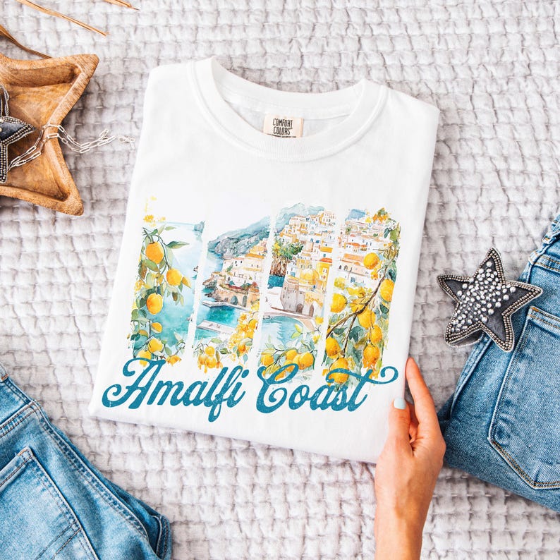 Pu&ograve; includere: Maglietta bianca con un'illustrazione ad acquerello della Costiera Amalfitana, Italia, con alberi di limone e il testo "Amalfi Coast" in blu.