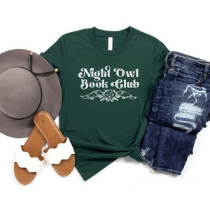 May include: A dark green t-shirt with the text "Night Owl Book Club" in white with a stylized owl graphic. The shirt is laid out on a white background with a hat, sandals, and jeans.