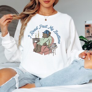 May include: White sweatshirt with a graphic of a bear wearing a blue sleep cap, sitting in a chair with a book and candles. The text "I Read Past My Bedtime" is written around the bear.