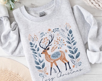 May include: A light gray sweatshirt featuring a deer design with floral accents in blue, orange, and brown. The deer has large antlers and is surrounded by leaves. The words "Cozy Christmas" are printed in a script font at the bottom.
