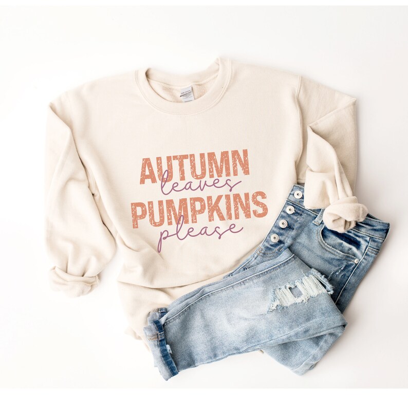 以下が含まれることがあります： オレンジと紫色の「AUTUMN leaves PUMPKINS please」の文字が入ったクリーム色のスウェットシャツ。ボタンのディテールとダメージ加工のパッチが付いたライトブルーのデニムジーンズがスウェットシャツの横に折りたたまれています。画像は白い背景にあります。