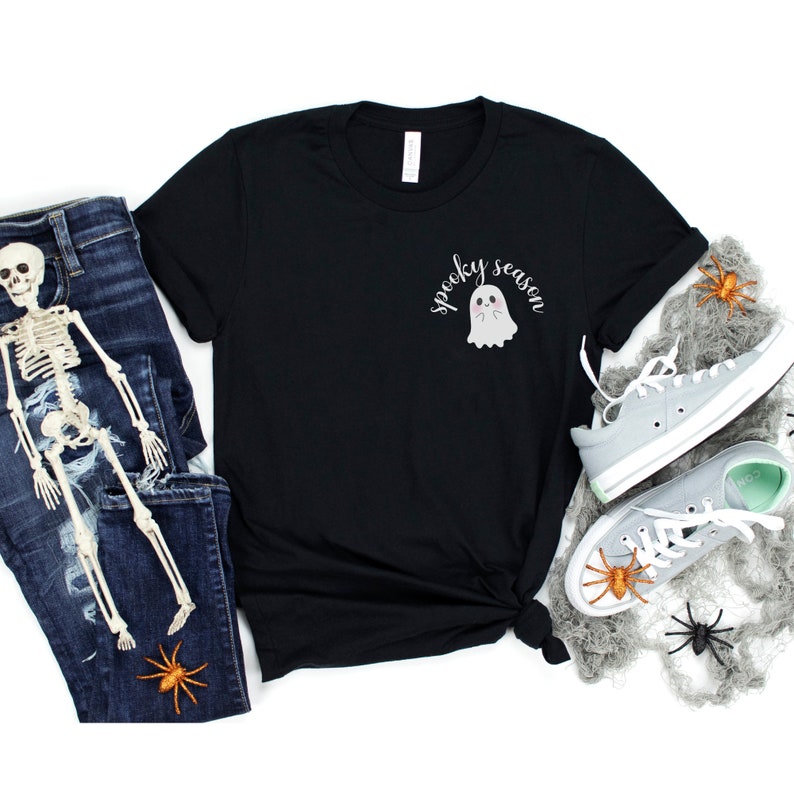 May include: Black t-shirt with a white ghost graphic and the text "spooky season". The shirt is laid out on a white background with a pair of gray sneakers, a skeleton, and spiders.