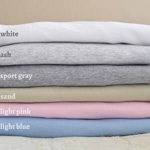 May include: Stack of six t-shirts in different colors: white, ash, sport gray, sand, light pink, and light blue.