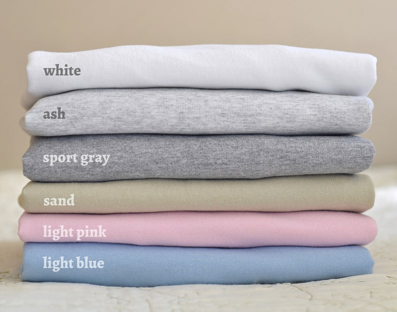 May include: Stack of six t-shirts in different colors: white, ash, sport gray, sand, light pink, and light blue.