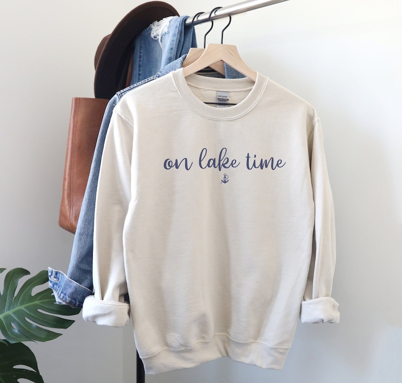 Puede incluir: Una sudadera de color crema con el texto "on lake time" en letra azul con un peque&ntilde;o s&iacute;mbolo de ancla debajo del texto.