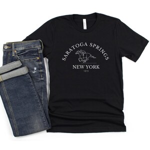 Puede incluir: Camiseta negra con un dise&ntilde;o gr&aacute;fico blanco de un caballo y un jinete. El gr&aacute;fico dice "Saratoga Springs New York 1819".