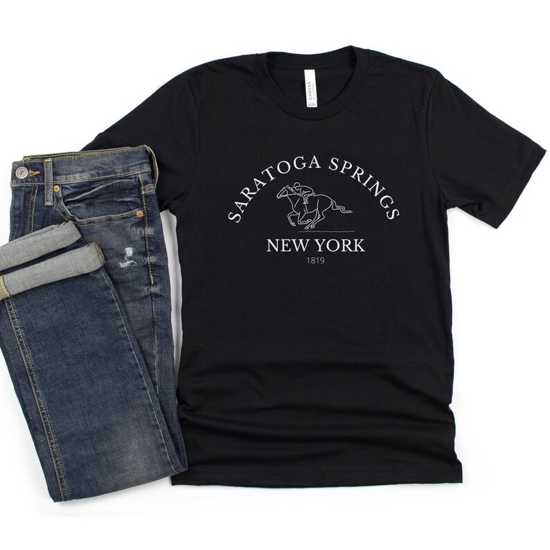 Puede incluir: Camiseta negra con un dise&ntilde;o gr&aacute;fico blanco de un caballo y un jinete. El gr&aacute;fico dice "Saratoga Springs New York 1819".