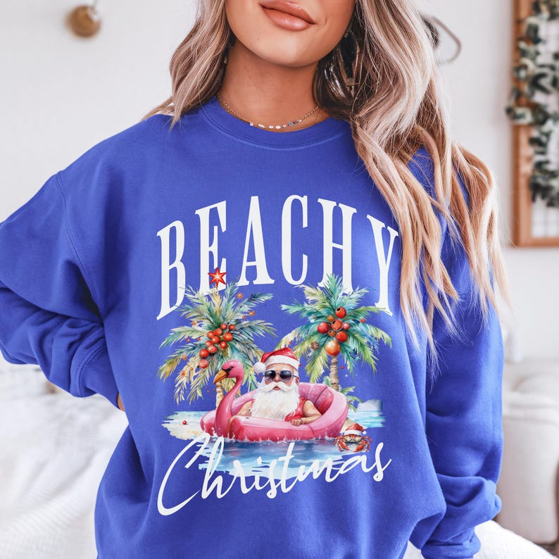 May include: Royal blue sweatshirt with a graphic featuring Santa Claus in sunglasses relaxing on a pink flamingo float in the ocean. The words "BEACHY Christmas" are printed above and below the image.