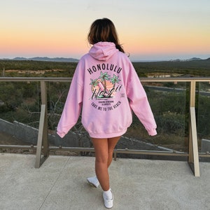May include: Pink hoodie with a graphic print of palm trees and surfboards. The text on the hoodie says "Honolulu" and "Take me to the beach".