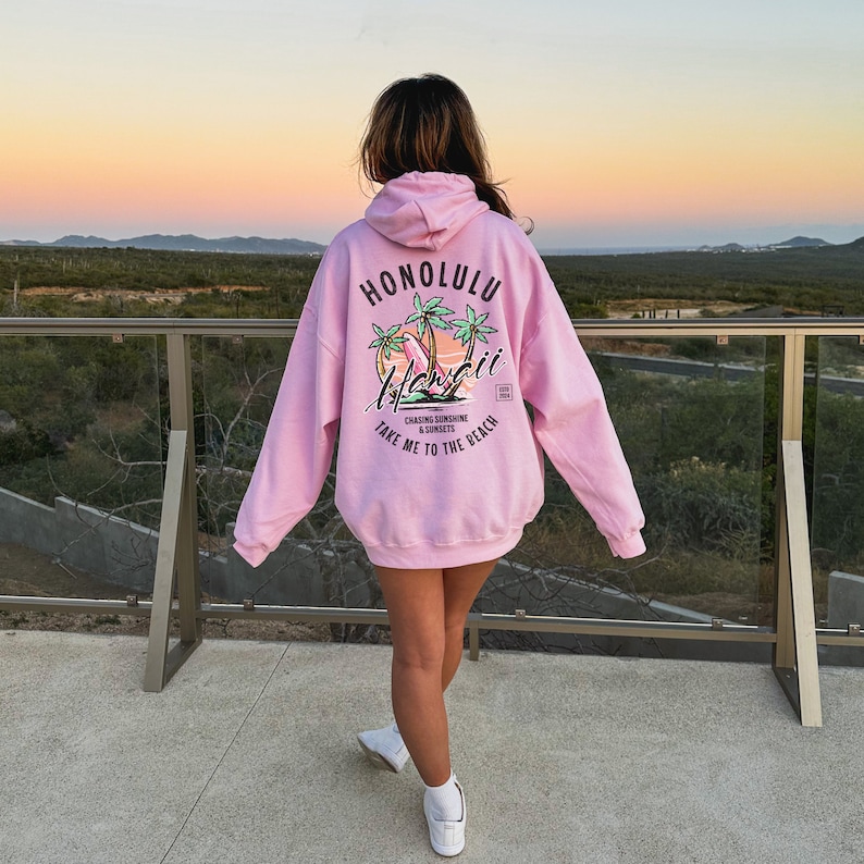 May include: Pink hoodie with a graphic print of palm trees and surfboards. The text on the hoodie says "Honolulu" and "Take me to the beach".