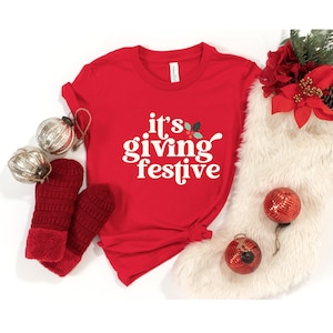 May include: A red t-shirt with the phrase "it's giving festive" in white script. The shirt is styled with red mittens, silver ornaments, and Christmas decorations, creating a festive holiday theme.