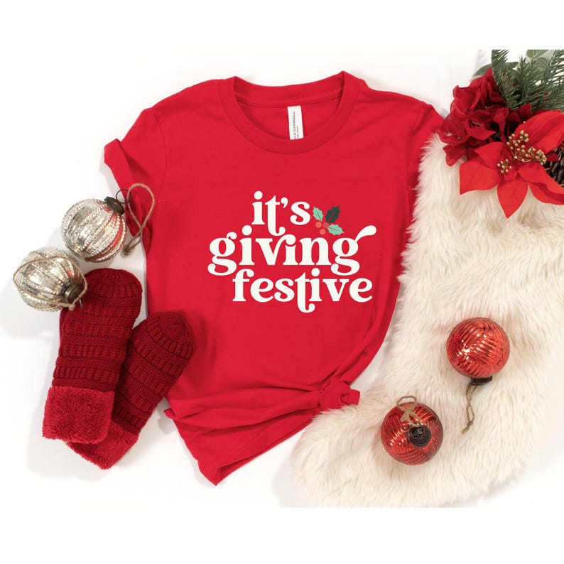 May include: A red t-shirt with the phrase "it's giving festive" in white script. The shirt is styled with red mittens, silver ornaments, and Christmas decorations, creating a festive holiday theme.