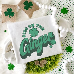 K&ouml;nnte beinhalten: Wei&szlig;es Langarm-Sweatshirt mit dem Schriftzug "Dibs on the Ginger" in Gr&uuml;n und einem Kleeblatt-Motiv. Das Design hat ein St. Patrick's Day-Thema. Kleeblatt-Dekorationen und ein Jutef&auml;hnchen sind im Hintergrund.