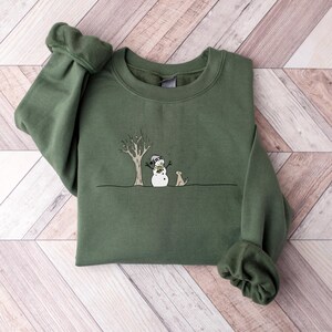 Op de afbeelding: Een olijfgroene sweatshirt met een grillige illustratie van een sneeuwpop, een hond en een kale boom. De sneeuwpop draagt een hoed en een sjaal. De hond zit naast de sneeuwpop. Het ontwerp is in zwart en wit.