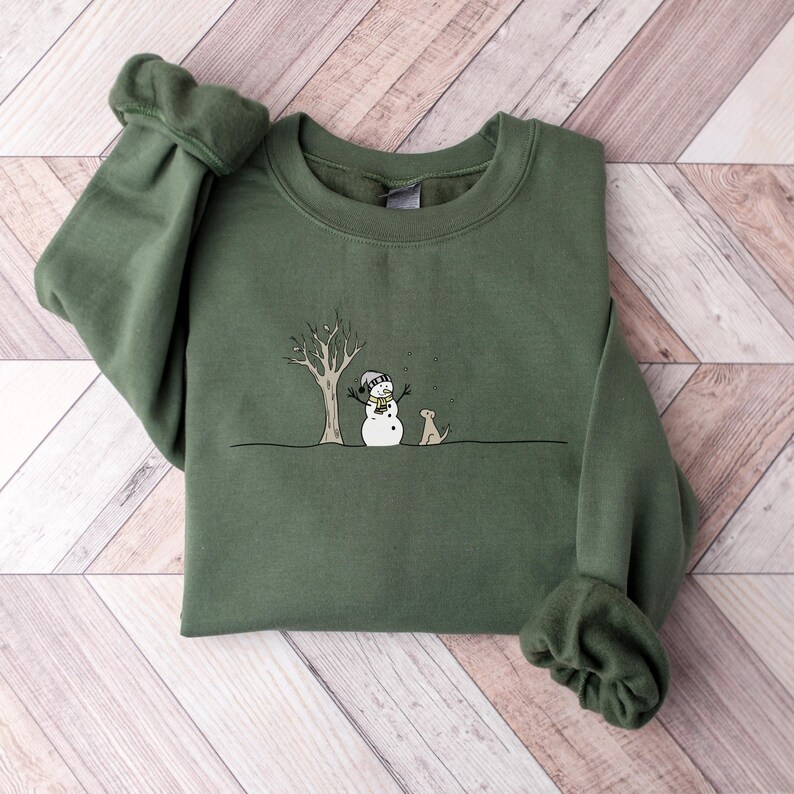 Op de afbeelding: Een olijfgroene sweatshirt met een grillige illustratie van een sneeuwpop, een hond en een kale boom. De sneeuwpop draagt een hoed en een sjaal. De hond zit naast de sneeuwpop. Het ontwerp is in zwart en wit.