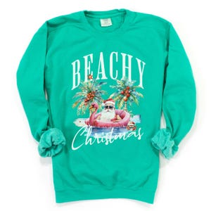 May include: Teal sweatshirt with a graphic of Santa Claus in sunglasses, floating on a pink flamingo in the ocean. Palm trees and the words "BEACHY Christmas" are above and below the illustration.